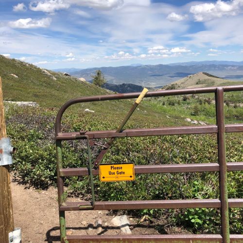 A metal gate on a hillside with a sign that says "Please Use Gate," overlooking a scenic valley and mountains.