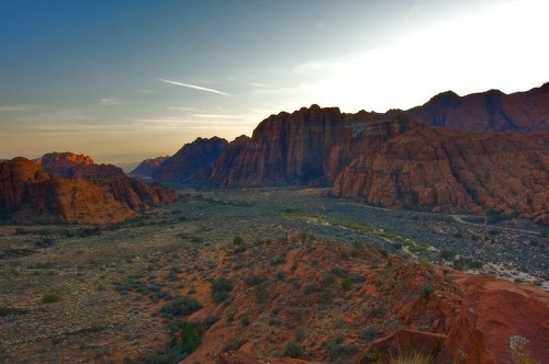 A scenic view of rugged red rock mountains at sunset, with a valley and river winding through the landscape.