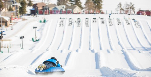 A person sledding down a snowy hill on a blue tube, with multiple lanes and winter scenery in the background.
