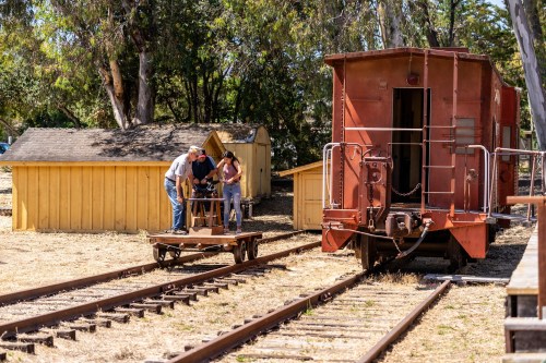 Two people work on a wooden cart near a red train car and yellow buildings in a sunny outdoor setting.