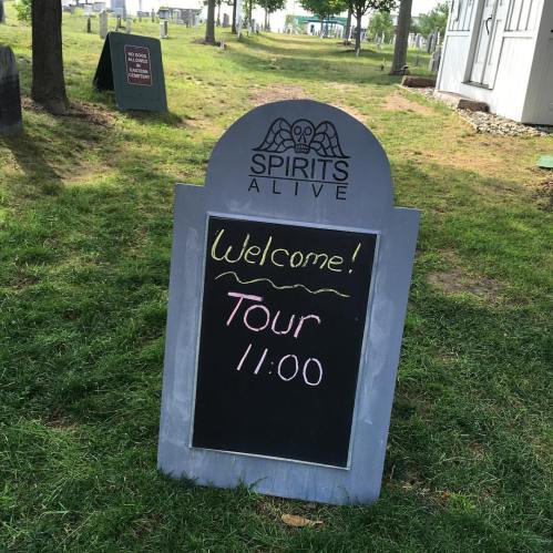 A chalkboard sign in a cemetery reads "Welcome! Tour 11:00" with a logo for "Spirits Alive" above it.