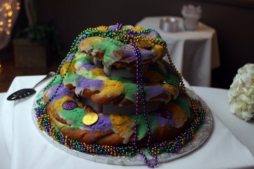A colorful king cake stacked on a silver platter, adorned with beads and gold coins, set on a white table.