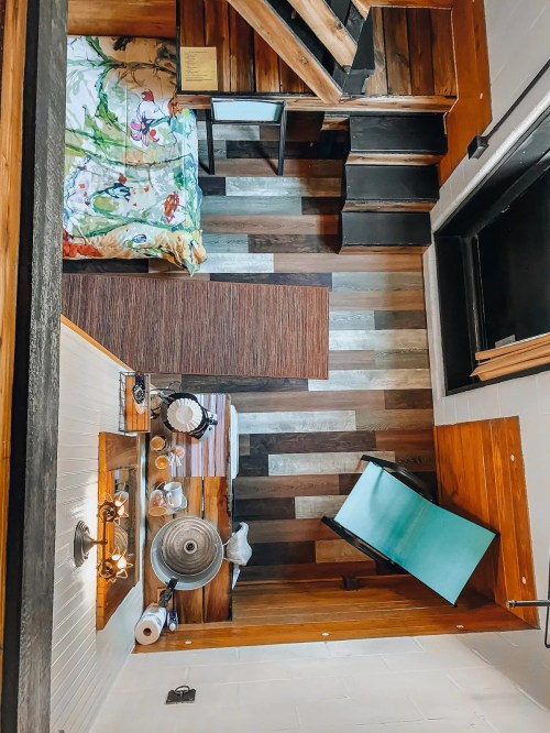 A cozy, wooden interior with a bed, stairs, and a small dining area, featuring colorful bedding and warm lighting.