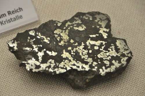 A metallic mineral specimen with a textured surface, displaying shiny silver patterns against a dark background.