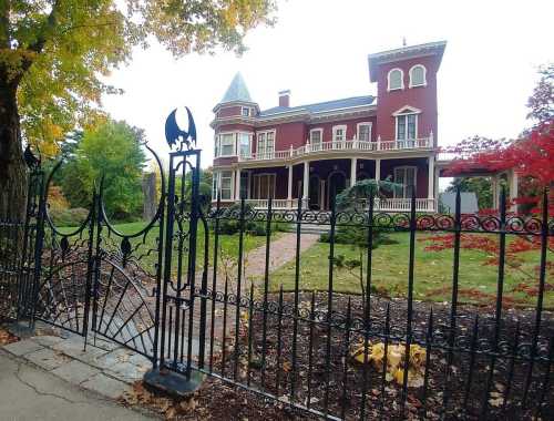 A large, red Victorian mansion with a tower, surrounded by a wrought iron fence and colorful autumn foliage.