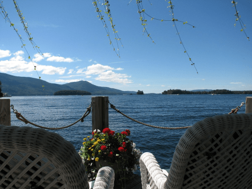A serene lakeside view with wicker chairs, flowers, and mountains under a clear blue sky.