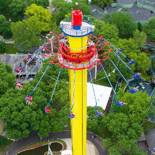 Aerial view of a tall amusement park ride with colorful swings and riders enjoying the thrill among green trees.