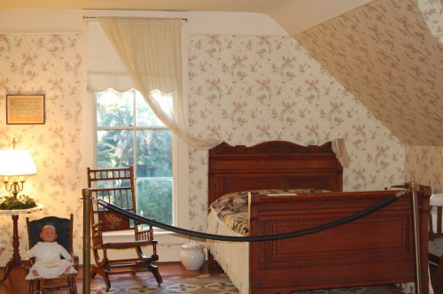 A vintage bedroom with floral wallpaper, a wooden bed, rocking chairs, and a doll on a chair. Natural light from a window.