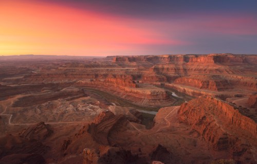 A stunning sunset over a vast canyon landscape, with layered rock formations and a winding river below.