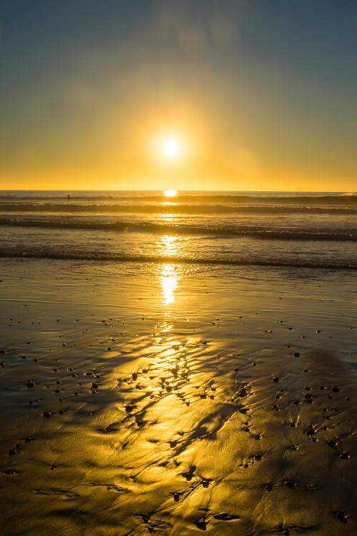 Sunset over a calm beach, with golden reflections on wet sand and gentle waves lapping at the shore.