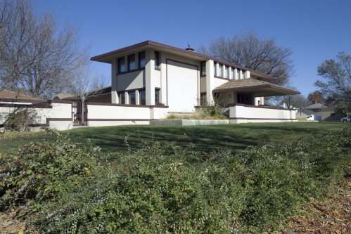 A modernist house with a flat roof, large windows, and a landscaped yard, surrounded by trees and shrubs.