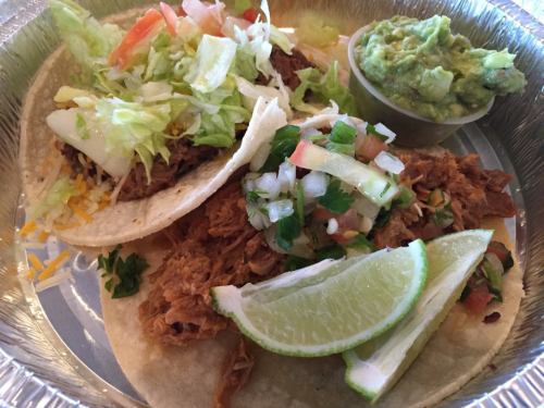 Two tacos filled with shredded meat, topped with lettuce and pico de gallo, served with guacamole and lime wedges.