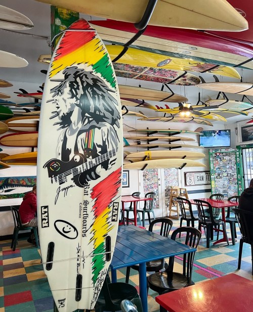 A colorful surfboard with a reggae design hangs in a vibrant surf-themed café with surfboards on the ceiling.