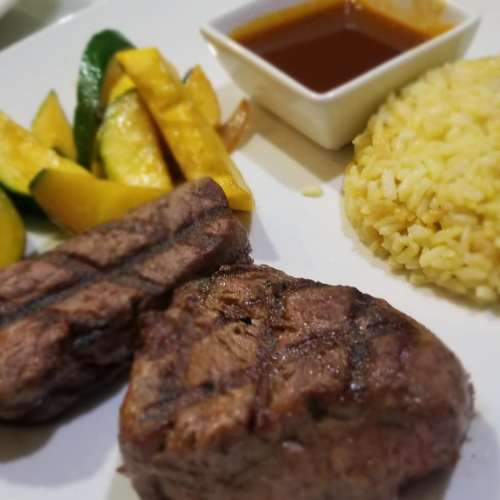 Grilled steak with sautéed zucchini, yellow squash, and a side of rice, accompanied by a small bowl of sauce.