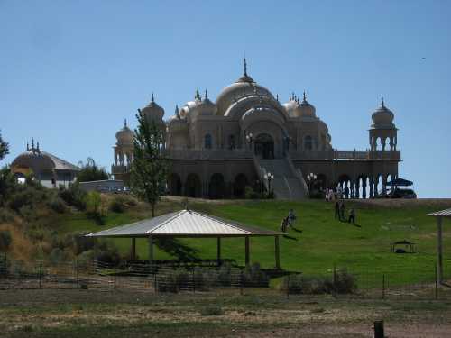 A grand, ornate building with domes and spires, set on a grassy hill with people walking nearby. Clear blue sky above.