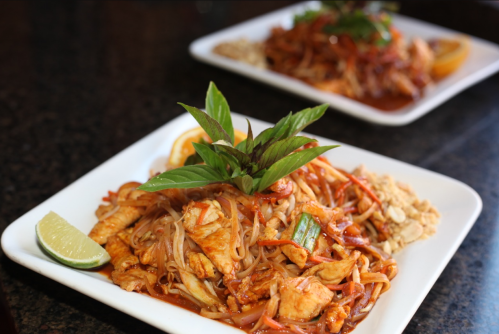 Two plates of colorful stir-fried noodles with chicken, vegetables, lime, and fresh herbs on a dark countertop.