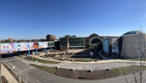 A panoramic view of a large building with colorful billboards, surrounded by greenery and a clear blue sky.