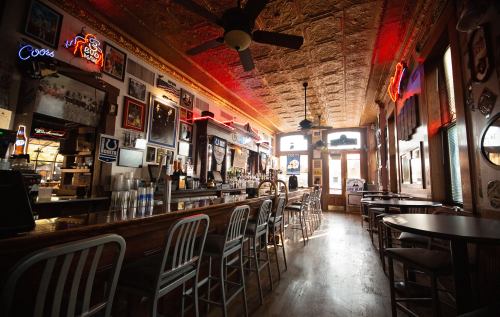 A cozy bar interior with a wooden floor, neon lights, and a long counter lined with stools.