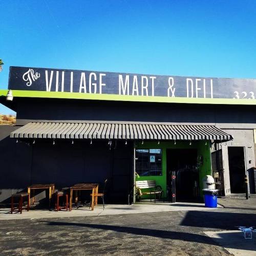 Exterior of The Village Mart & Deli, featuring a green entrance and outdoor seating under a striped awning.