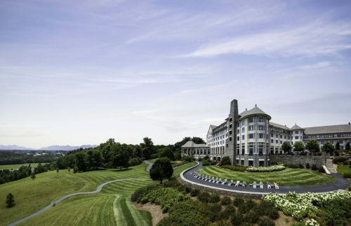 A scenic view of a large stone hotel surrounded by lush green lawns and mountains in the background under a clear sky.