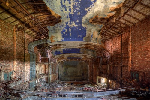 Abandoned theater interior with crumbling walls, a damaged ceiling, and remnants of a stage, evoking a sense of decay.