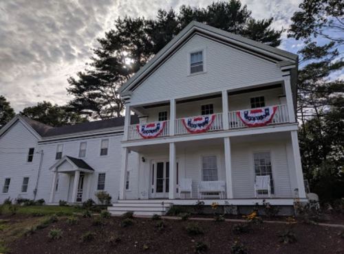 A white two-story house with red, white, and blue bunting on the balconies, surrounded by greenery and cloudy skies.