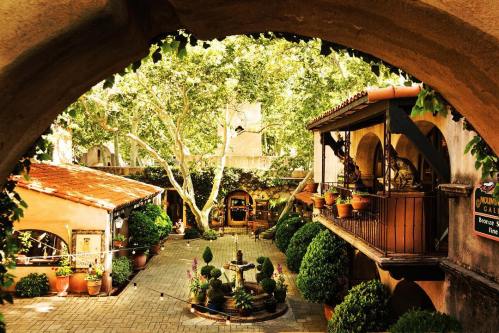 A charming courtyard with lush greenery, potted plants, and a fountain, framed by archways and rustic buildings.