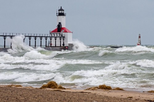 A lighthouse stands on a pier as waves crash against it, with a cloudy sky in the background.