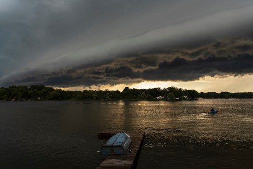 A stormy sky looms over a calm lake, with dark clouds and a lone boat on a wooden dock.