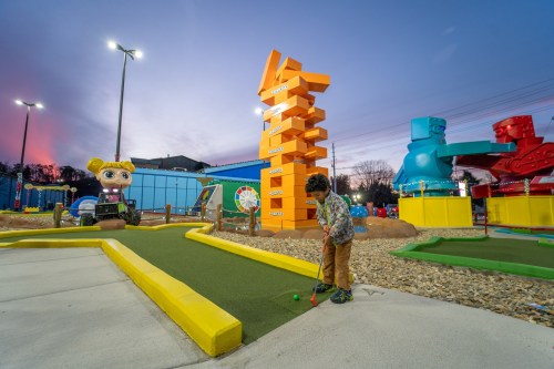 A child plays mini-golf at sunset, with colorful sculptures and a large Jenga tower in the background.