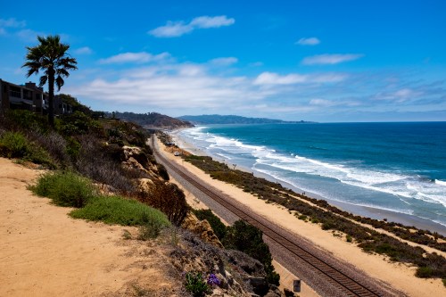 A scenic coastal view with sandy cliffs, railway tracks, and waves crashing on the shore under a blue sky.