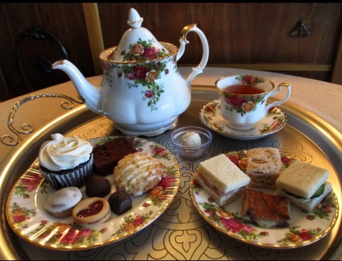 A floral teapot and cup with tea, surrounded by plates of assorted pastries, sandwiches, and desserts on a tray.