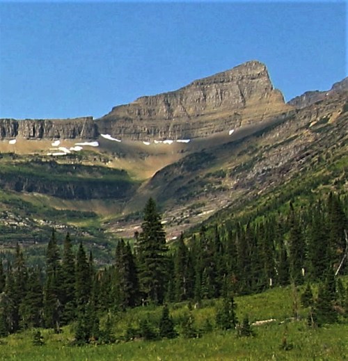 A scenic mountain landscape featuring a prominent peak, lush green trees, and a clear blue sky.