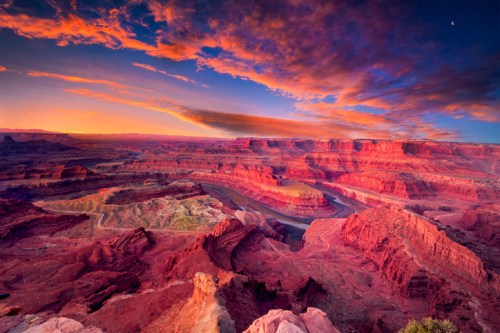 Vibrant sunset over a vast canyon, with layered red rock formations and a winding river below, under a colorful sky.