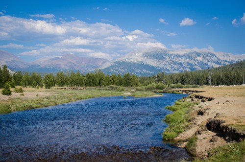 A serene river flows through a lush landscape, surrounded by mountains and blue skies with scattered clouds.