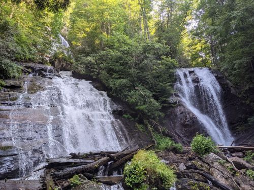 A serene waterfall cascades down rocky cliffs, surrounded by lush green trees and fallen logs in a tranquil forest setting.
