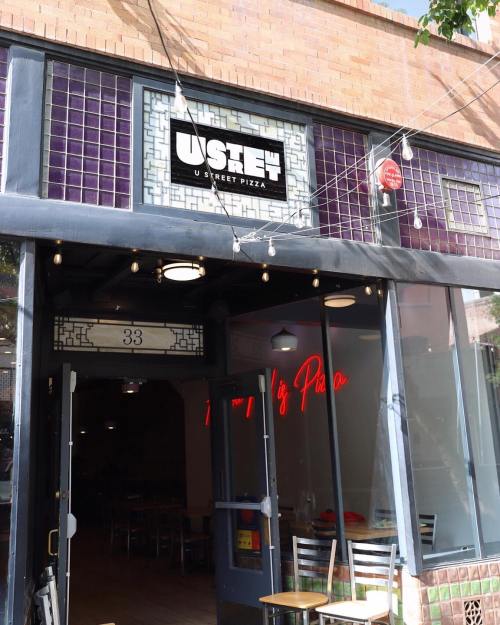 Entrance of U Street Pizza, featuring a sign and neon lights, with outdoor seating visible.