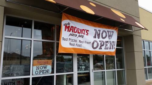 A storefront with a large banner reading "Uncle Maddio's Pizza Joint - Now Open!" promoting fresh pizza.