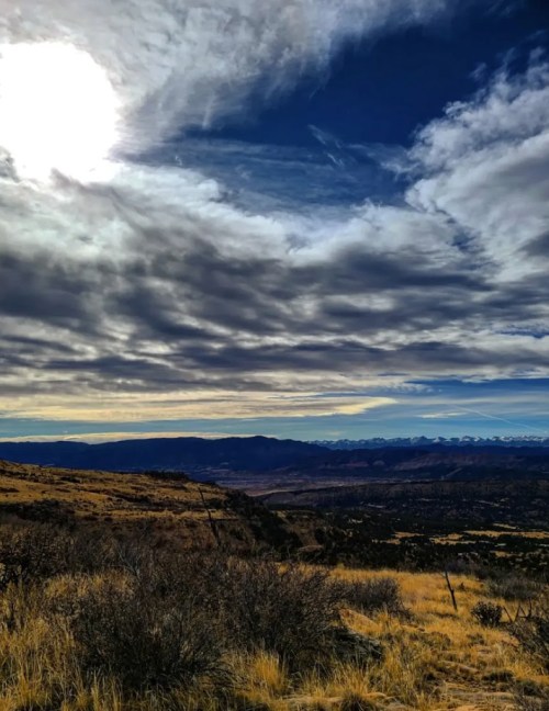 A scenic view of rolling hills under a cloudy sky, with distant mountains and a bright sun illuminating the landscape.