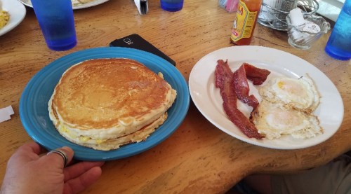 A plate with large pancakes and a side of bacon and two fried eggs on a wooden table.