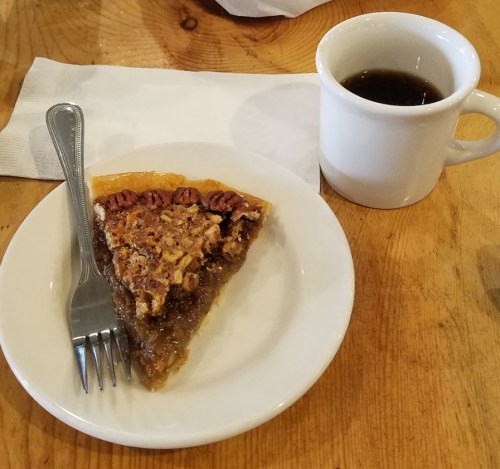 A slice of pecan pie on a white plate with a fork, next to a cup of black coffee on a wooden table.