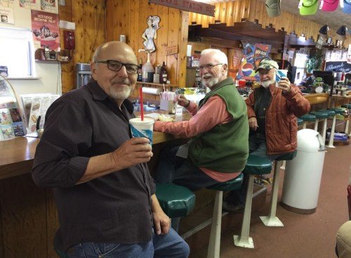 Three men sit at a diner counter, smiling and holding drinks, with a cozy, retro atmosphere in the background.