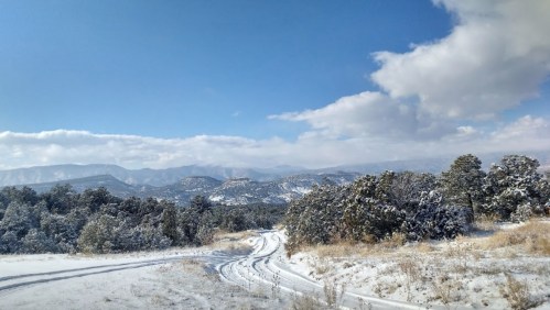 A snowy landscape with winding paths, trees, and mountains under a blue sky with scattered clouds.