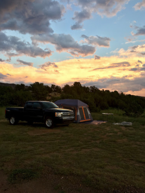A black pickup truck parked next to a tent under a colorful sunset with clouds in a grassy campsite.