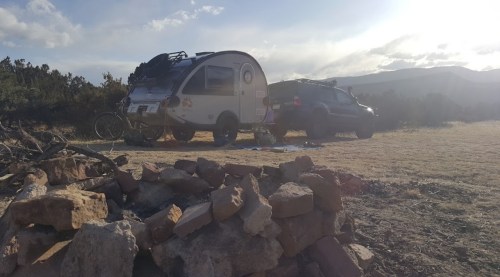 A small camper and an SUV parked in a desert landscape, with a rock pile in the foreground and a cloudy sky.