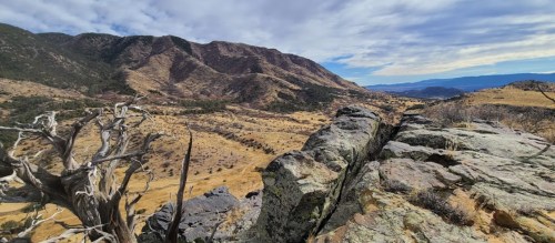 A panoramic view of rolling hills and mountains under a cloudy sky, with rocky terrain in the foreground.