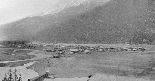 A black-and-white landscape of a small village by a river, surrounded by mountains and trees.