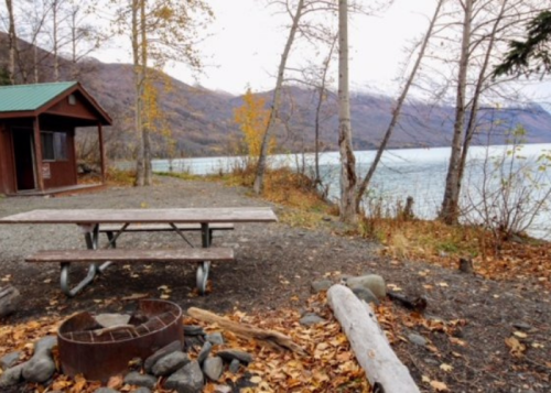 A serene lakeside scene with a cabin, picnic table, fire pit, and autumn foliage against a mountain backdrop.