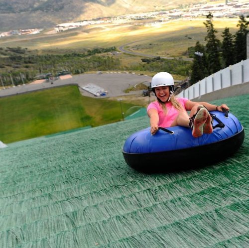 A person in a helmet joyfully sleds down a green slope, surrounded by scenic hills and a clear sky.