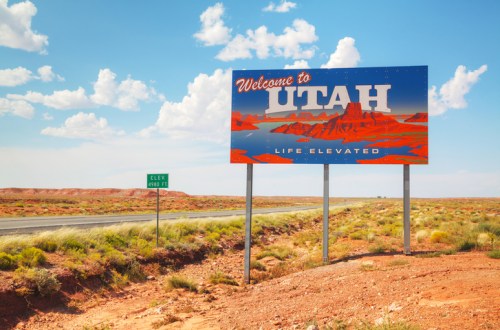 A colorful "Welcome to Utah" sign stands beside a highway, with a blue sky and desert landscape in the background.